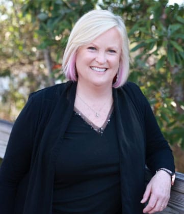 Woman with white-blonde hair smiling at camera, wearing black cardigan over black top, outdoor foliage background.