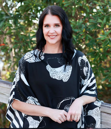 Woman with long dark hair smiling at camera wearing patterned black and white top against green foliage backdrop.