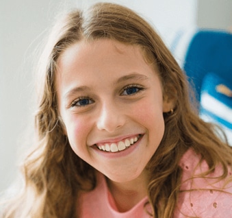 Young girl with long brown hair smiling at camera, wearing pink shirt, indoors.
