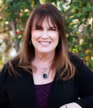 Woman with brown hair and bangs smiling at camera against blurred green foliage background.