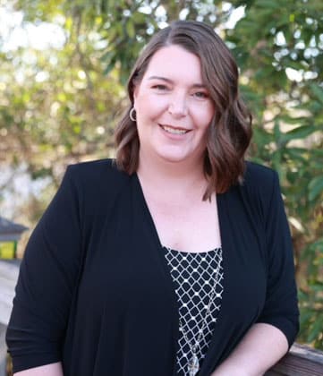 Woman smiling at camera wearing black cardigan over patterned top, outdoors with blurred green foliage background.