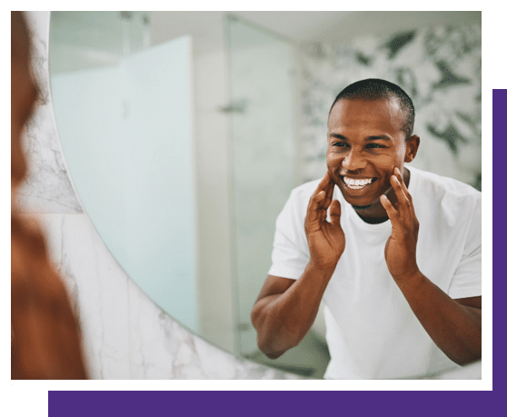 Man in white shirt smiling at bathroom mirror while touching his face.