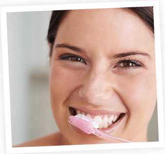 Woman brushing teeth with pink toothbrush, smiling at camera indoors.