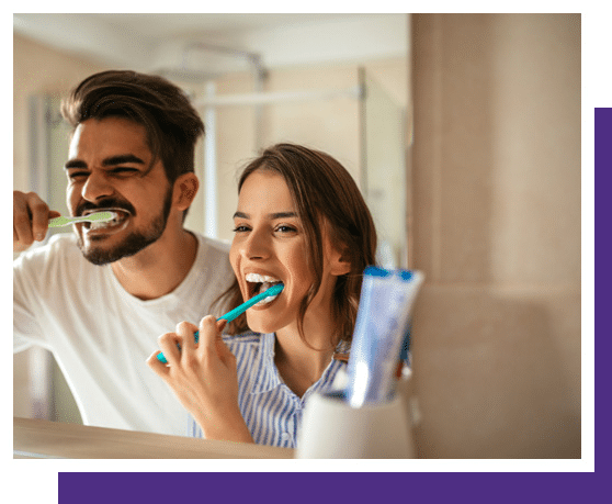Young couple brushing teeth together in bathroom mirror, both smiling.