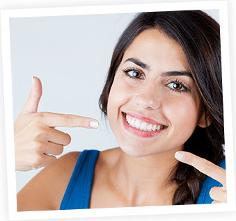 Smiling woman pointing at her teeth against white background, wearing blue top.