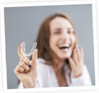 Woman holding clear dental retainer, smiling happily against gray background.