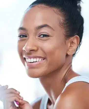 Young woman smiling at camera, holding skincare product, bright indoor setting.