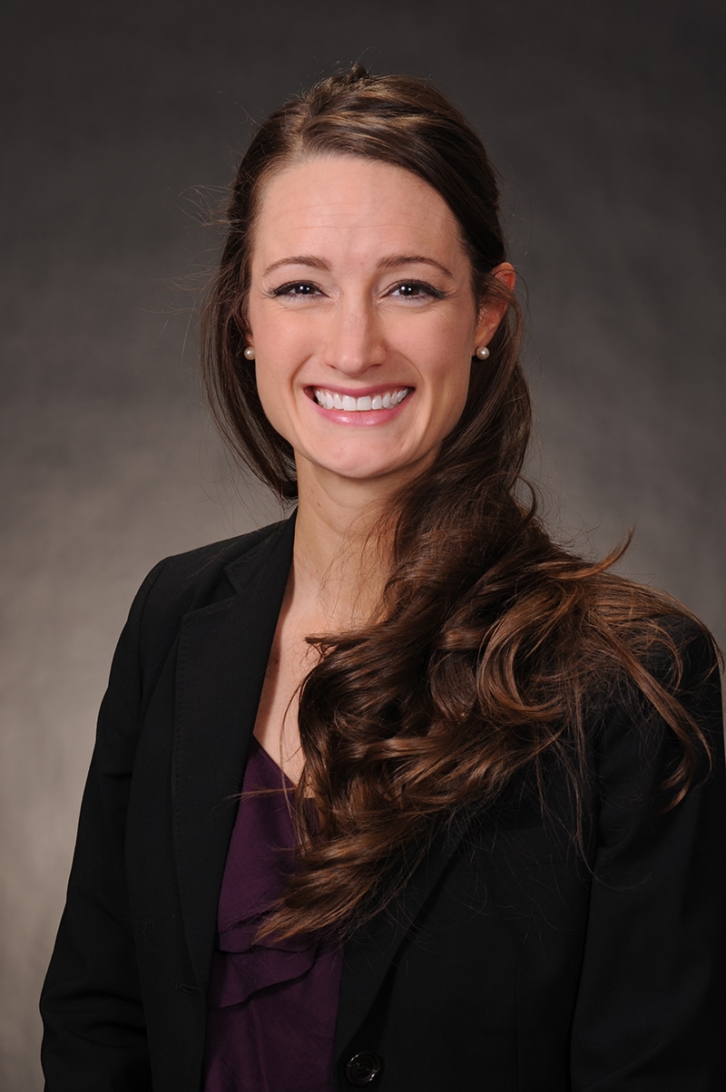 Professional headshot of a smiling woman with long brown hair wearing a black blazer and purple top against a gray background.