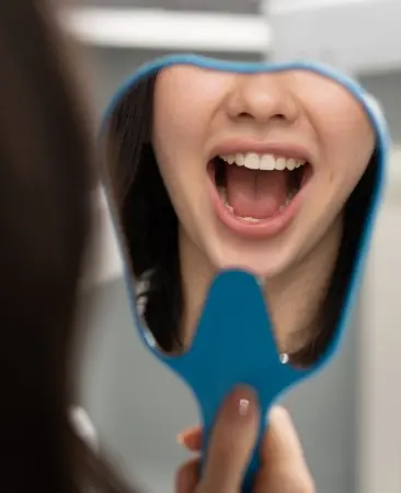 Woman smiling brightly while holding a blue handheld mirror showing her healthy teeth.