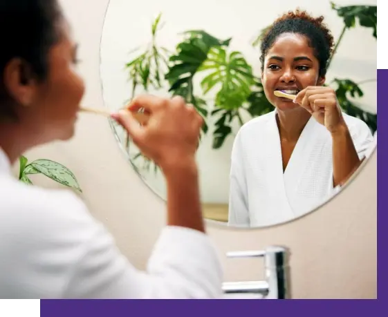 Woman brushing teeth in bathroom mirror with green plants, representing holistic dental care.